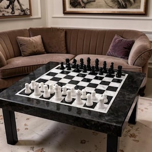 Könnte beinhalten: Ein quadratischer Schach-Tisch aus schwarz-weißem Marmor mit einem kompletten Schachspiel. Die Schachfiguren sind schwarz und weiß. Der Tisch hat schwarze Beine. Im Hintergrund ein braunes Sofa mit Kissen.
