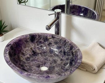 May include: A round, purple amethyst stone sink with a polished finish. The sink is set on a white countertop with a chrome faucet. A folded beige towel rests on the counter. The sink is reflected in a mirror.