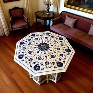 May include: An octagonal coffee table with a white marble top inlaid with blue and gold floral patterns. The table has a decorative base and sits on a hardwood floor. The table's dimensions are approximately 48 inches in diameter.