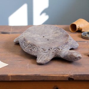 Puede incluir: Jabonera gris con forma de tortuga. El plato tiene un cuenco poco profundo con dos agujeros de drenaje y tallas decorativas. La cabeza y las aletas de la tortuga est&aacute;n detalladas.