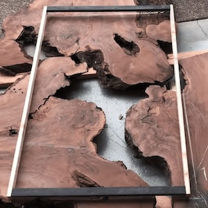 May include: Unfinished wooden slabs with natural edges and a dark reddish-brown hue. A rectangular frame sits on one slab, and a measuring stick is present, indicating woodworking or furniture construction. The wood is likely for a table.