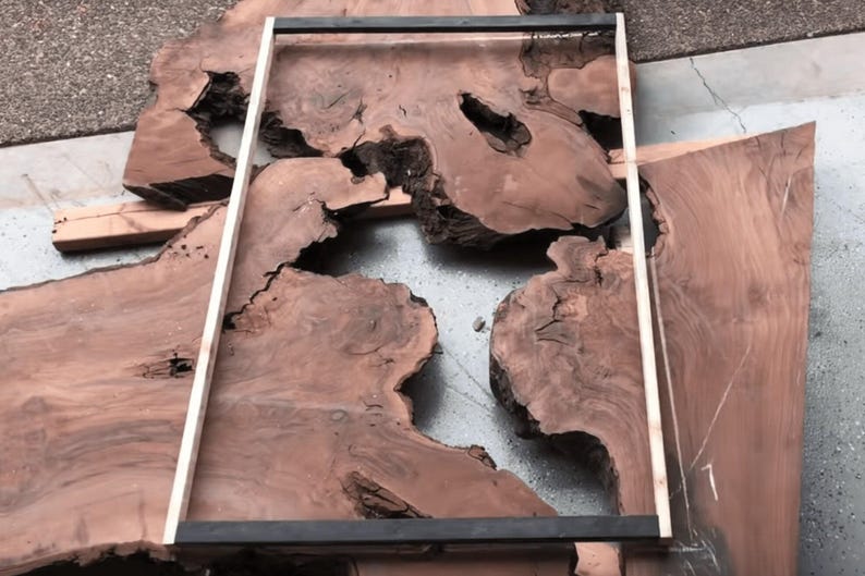 May include: Unfinished wooden slabs with natural edges and a dark reddish-brown hue. A rectangular frame sits on one slab, and a measuring stick is present, indicating woodworking or furniture construction. The wood is likely for a table.