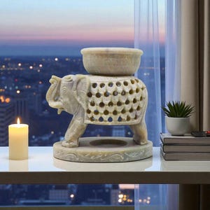 May include: A carved stone elephant incense burner with a bowl on top. The elephant has a decorative, openwork design on its back. A lit candle and a small potted plant are also visible.