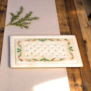 May include: Rectangular white marble tray with a decorative floral border in green, red, and brown. The tray has a textured surface with a repeating pattern. The tray is on a light gray surface and a wooden table.