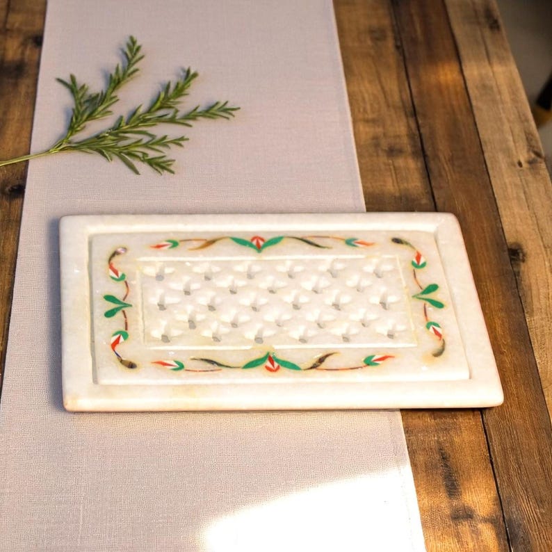 May include: Rectangular white marble tray with a decorative floral border in green, red, and brown. The tray has a textured surface with a repeating pattern. The tray is on a light gray surface and a wooden table.