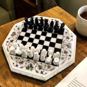 Könnte beinhalten: Ein dekoratives Schachspiel mit schwarzen und weißen Figuren auf einem Schachbrett. Das Brett ist weiß mit floralen Akzenten und steht auf einem Holztisch. Eine Tasse Tee und ein Buch befinden sich ebenfalls auf dem Tisch.