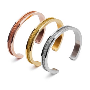 May include: Three minimalist bracelets in rose gold, gold, and silver with a textured, sparkly finish.