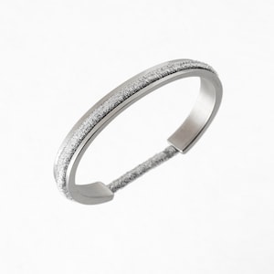 May include: A silver bangle bracelet with a textured, glittery band. The bracelet features a simple, modern design with a smooth, polished finish on the outer edges. The textured band adds a touch of sparkle.