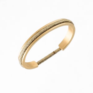 May include: A gold-toned metal bracelet with a textured band. The band is made of a woven material with a subtle shimmer.