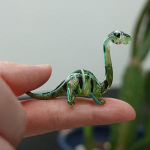 Może przedstawiać: Miniaturowa szklana figurka dinozaura. Figurka jest głównie zielona z czarnymi i złotymi akcentami. Dinozaur ma długą szyję i ogon i jest umieszczony na palcu.