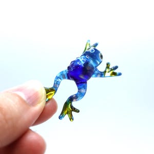 Puede incluir: Una pequeña figura de rana de cristal en tonos azules y amarillos. La rana tiene un cuerpo azul oscuro y patas azul claro con pies amarillos. La figura se sostiene en una mano sobre un fondo blanco.