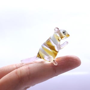 May include: A miniature glass squirrel figurine with a striped pattern in shades of amber, white, and clear. The small sculpture is posed upright, with its tiny paws raised, and is resting on a finger.