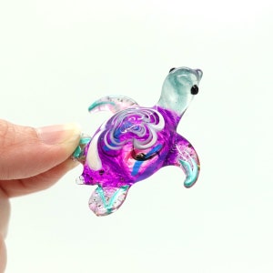 May include: A small, colourful glass turtle figurine. The turtle features a purple, white, and blue swirled design on its shell, with turquoise flippers and head. The figurine is held between two fingers against a white background.