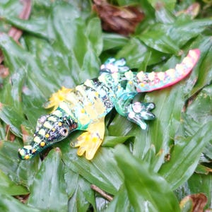May include: A colourful glass gecko figurine with green, yellow, pink and black accents. The gecko is posed on its belly with its legs outstretched. The figurine is sitting on a bed of green grass.
