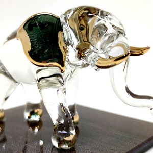 Op de afbeelding: Een transparant glazen olifantbeeldje met gouden accenten en een groen glazen oor. De olifant staat op een donkerbruine houten basis.