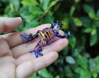 May include: A small glass frog figurine featuring a deep blue and purple translucent body with iridescent highlights. The frog has gold-coloured horizontal stripes across its back and detailed, splayed limbs with distinct toes, crafted in a smooth, glossy finish.