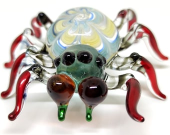 May include: A glass spider figurine with a colorful, swirled body and red legs. The spider has black eyes and a green abdomen.
