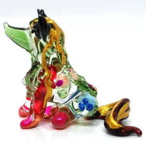 May include: A colorful glass sculpture of a dog with a curled tail. The dog is primarily clear with green, red, pink, blue, and gold accents. The sculpture has a whimsical, artistic design.