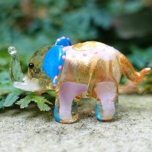 May include: A small glass elephant figurine with a pink body, gold accents, and blue ears. The elephant has a white trunk and is standing on a grey surface.