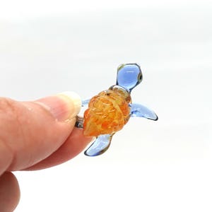May include: A small glass sea turtle figurine with a blue body and an orange shell. The turtle is hand-blown and has a unique, speckled texture.