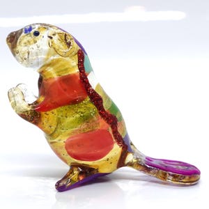 May include: A colourful glass figurine of a beaver with a purple tail, red, green, and yellow stripes, and gold glitter accents.