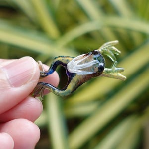 以下が含まれることがあります： 詳細なデザインの小さなガラス製のカエルフィギュア。カエルは、黄色、青、白などの色の組み合わせが特徴です。フィギュアは人の手で持たれており、そのサイズと複雑な職人技を示しています。