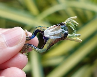 May include: A miniature glass frog ornament, showcasing a detailed design. The frog is painted with yellow, blue, and white hues. The figurine is held in a hand, highlighting its size and intricate craftsmanship.