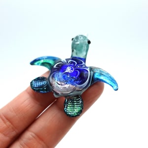 Puede incluir: Una pequeña figura de tortuga marina de vidrio con cabeza y aletas de color verde claro translúcido, con un patrón de remolinos azul profundo y blanco en el caparazón. El caparazón incluye acentos plateados brillantes y detalles verdes texturizados.