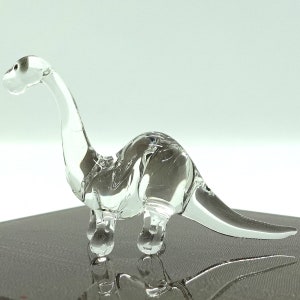 Może przedstawiać: Szklana figurka dinozaura wykonana z przezroczystego szkła, z długą szyją i ogonem. Figurka jest ozdobiona fakturowanym szkłem i ma lekko zamglony wygląd.