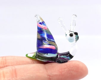 May include: A colorful glass snail figurine. The snail's shell is a vibrant mix of blue, pink, and gold, while its body is clear with black and purple accents. The figurine is held on a finger.