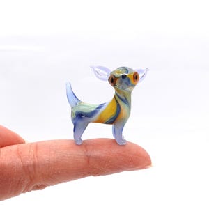 Peut inclure: Une petite figurine en verre d'un chien chihuahua. La figurine est bleue, jaune et blanche avec des motifs en spirale. Le chien a de grands yeux ronds.