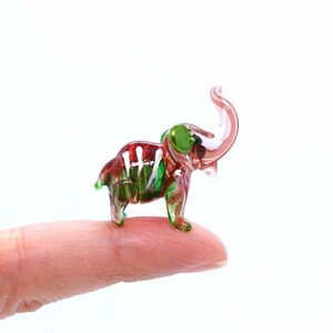 May include: A small glass elephant figurine with red, green, and white details. The elephant is standing on a finger.