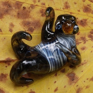以下が含まれることがあります： 黒と白のストライプのナマケモノのガラス彫刻で、黄色のガラスの目があります。彫刻は、茶色の斑点のある黄色の葉の上に横たわっています。