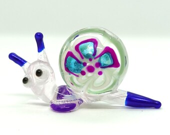 May include: A glass snail figurine with a white shell, blue antennae, and purple accents. The shell has a painted flower design with blue, pink, and white colors.