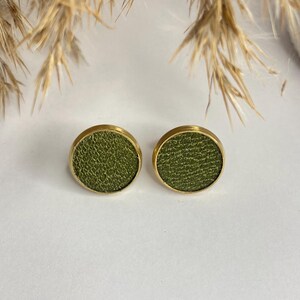 May include: A pair of round stud earrings. Each earring features a textured olive green center set in a gold-colored bezel. The earrings are displayed on a white surface, with dried ornamental grass in the background.