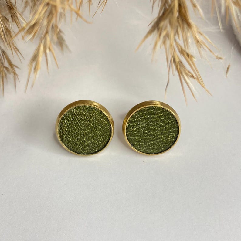 May include: A pair of round stud earrings. Each earring features a textured olive green center set in a gold-colored bezel. The earrings are displayed on a white surface, with dried ornamental grass in the background.