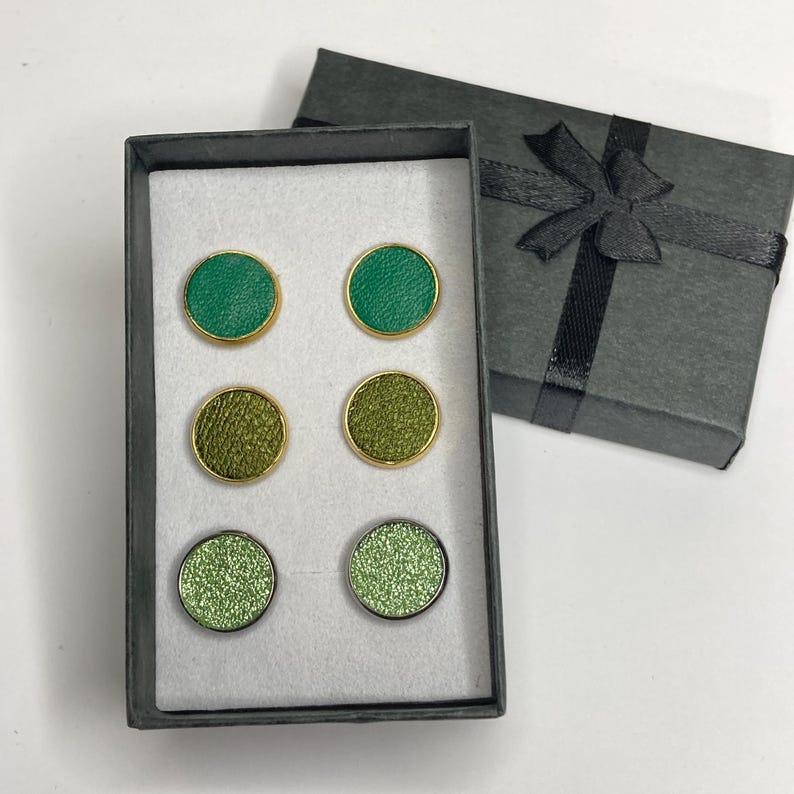 May include: A set of six green and gold cufflinks in a gray gift box. The cufflinks are arranged in pairs, with varying shades of green, including a glittery pair. The box is open, revealing the cufflinks, and a closed box with a black ribbon is next to it.