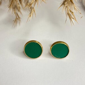 May include: A pair of round stud earrings. Each earring features a vibrant green center set in a gold-colored metal frame. The earrings are displayed on a white surface, with dried plants in the background.