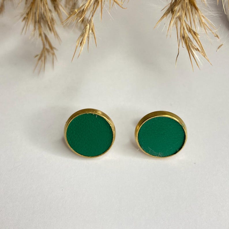 May include: A pair of round stud earrings. Each earring features a vibrant green center set in a gold-colored metal frame. The earrings are displayed on a white surface, with dried plants in the background.