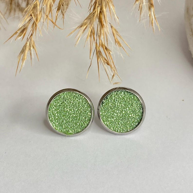 May include: A pair of round stud earrings with a light green glittery surface set in a silver-colored metal. The earrings are displayed on a white surface with dried ornamental grass in the background.