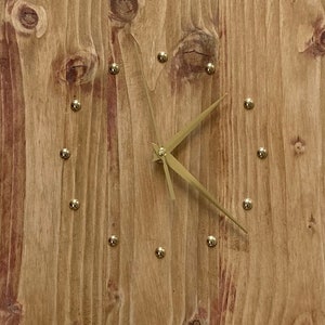Peut inclure: Une horloge en bois avec des aiguilles dorées et des clous dorés autour du bord. Le cadran de l'horloge est en bois naturel avec une finition rustique. L'horloge est signée "Créac'ile" dans le coin inférieur gauche.
