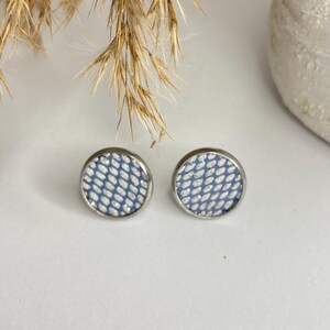 May include: A pair of round stud earrings. Each earring features a blue and white patterned design set within a silver-colored metal frame. The earrings are displayed on a white surface.