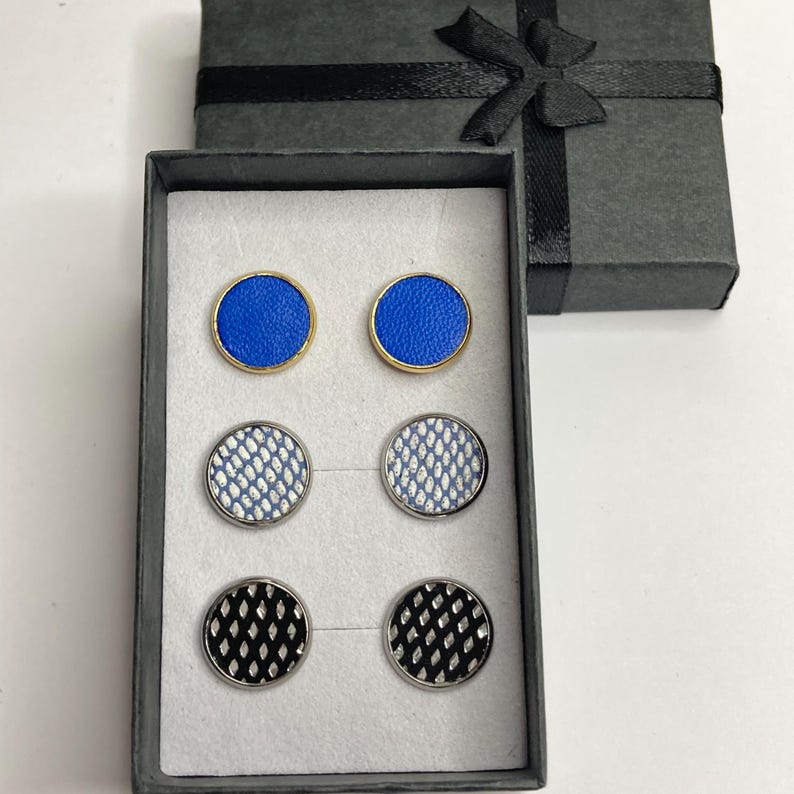 May include: A set of six cufflinks in a gray gift box. Two blue cufflinks with gold trim, two silver cufflinks with a white and blue pattern, and two black cufflinks with a silver pattern. The box is closed with a black ribbon.