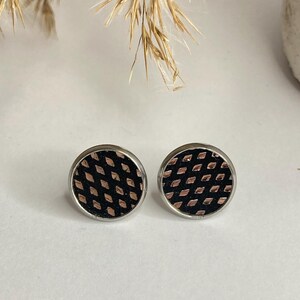 May include: A pair of round stud earrings. Each earring features a black background with a copper-colored diamond pattern, set within a silver-colored metal frame. The earrings are displayed on a white surface.