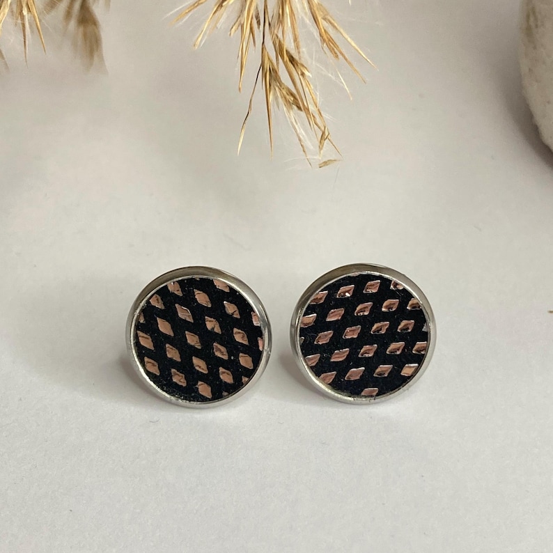 May include: A pair of round stud earrings. Each earring features a black background with a copper-colored diamond pattern, set within a silver-colored metal frame. The earrings are displayed on a white surface.