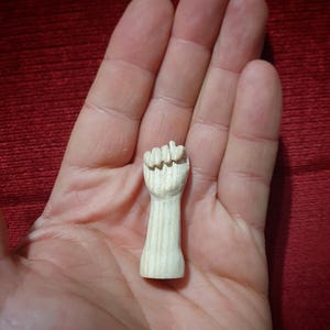 May include: A small, carved wooden fist figurine. The fist is clenched, with detailed finger and knuckle definition. The wood has a natural, light color and visible grain. The figurine is held in an open hand against a red background.