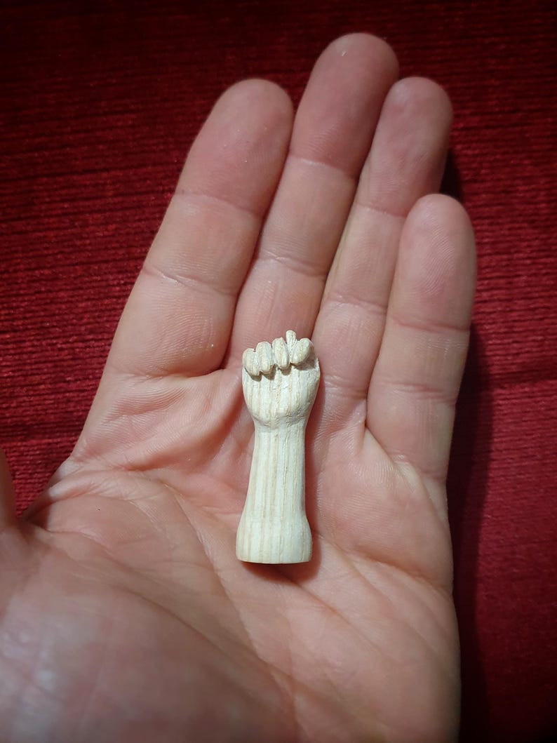 May include: A small, carved wooden fist figurine. The fist is clenched, with detailed finger and knuckle definition. The wood has a natural, light color and visible grain. The figurine is held in an open hand against a red background.