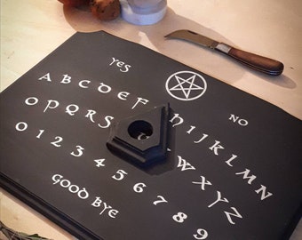 Könnte beinhalten: Schwarzes Ouija-Brett aus Holz mit weißen Buchstaben, einschließlich Alphabet, Zahlen 0-9, "ja", "nein" und "auf Wiedersehen". In der Mitte des Bretts befindet sich ein Pentagramm.