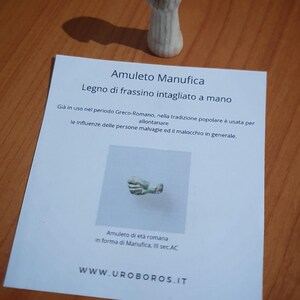 May include: A carved wooden amulet in the shape of a fist. The amulet is on a wooden surface with a printed card that reads "Amuleto Manufica" and "Legno di frassino intagliato a mano". The text also mentions its use in the Greco-Roman period.