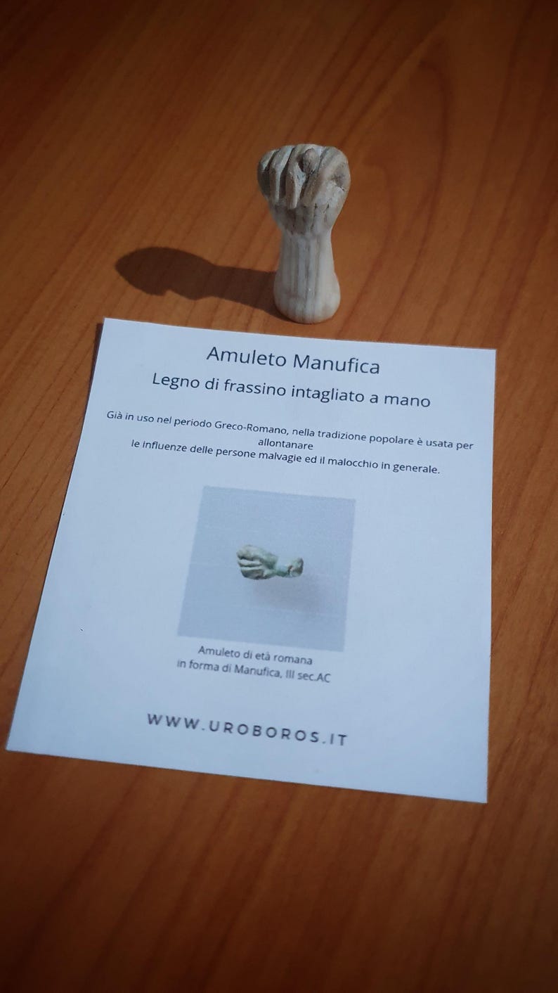 May include: A carved wooden amulet in the shape of a fist. The amulet is on a wooden surface with a printed card that reads "Amuleto Manufica" and "Legno di frassino intagliato a mano". The text also mentions its use in the Greco-Roman period.